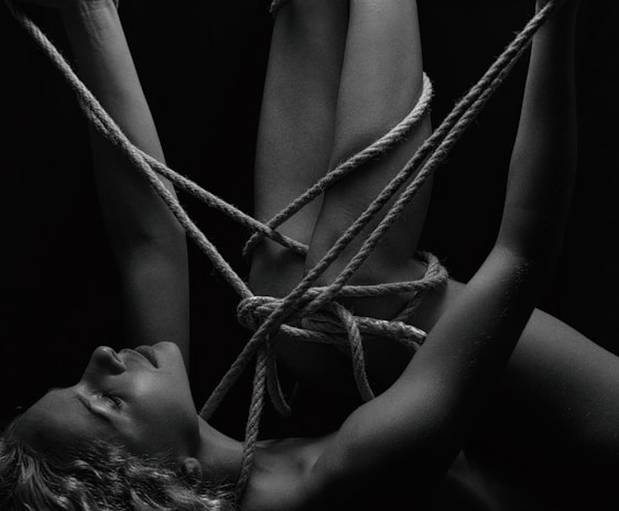 A person is intricately bound with ropes, creating a complex pattern around their body. The monochrome setting adds a stark contrast, emphasizing the textures of the skin and ropes. The person's eyes are closed, conveying a sense of calm or introspection.