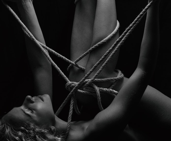A person is intricately bound with ropes, creating a complex pattern around their body. The monochrome setting adds a stark contrast, emphasizing the textures of the skin and ropes. The person's eyes are closed, conveying a sense of calm or introspection.