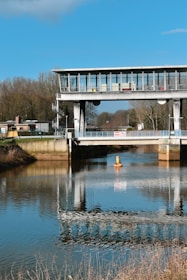 A modern bridge structure spans across a calm, reflective body of water. The bridge, made of metal and glass, features a control room with large windows. The blue sky and surrounding bare trees are mirrored almost perfectly on the water's surface.