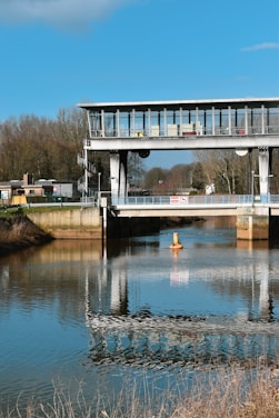 A modern bridge structure spans across a calm, reflective body of water. The bridge, made of metal and glass, features a control room with large windows. The blue sky and surrounding bare trees are mirrored almost perfectly on the water's surface.