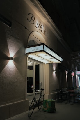 A night scene featuring the entrance of a café-bar-restaurant named Bellaria. The building exterior is illuminated by wall-mounted lamps casting light and shadow patterns on the textured walls. A classic bicycle is parked in front of the entrance, next to a green delivery bag. The sign above the door is well-lit, displaying the name and type of establishment.