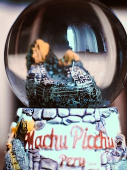 A glass globe containing a miniature model of Machu Picchu sits atop a base with artistic representations of stone and foliage. The text 'Machu Picchu, Peru' is prominently displayed on the base. Inside the globe, detailed features of the iconic Incan site are depicted in miniature, with hints of mountains and terraces.