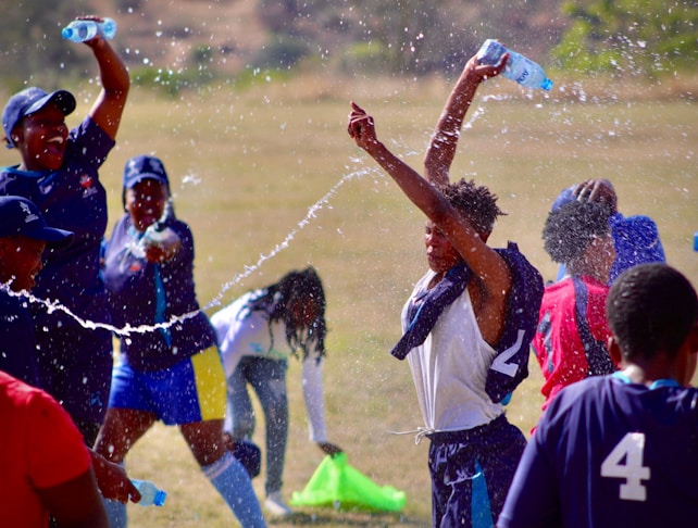 A group of people, mostly young, are joyfully spraying water at each other in a playful and energetic manner. They are outdoors on a field, and some are wearing sports jerseys or athletic clothing. The water is splashing through the air, creating a lively scene.