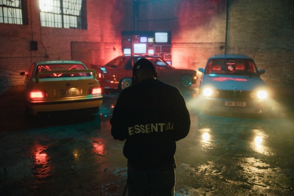 A dimly lit garage with three parked cars, each illuminated by colorful lighting. There is a person in the foreground wearing a jacket with the word 'ESSENTIAL' printed on the back. The setting has an industrial vibe with exposed brick walls, and several static-filled televisions mounted in the background. The lights create a moody atmosphere with reflections on the wet floor.