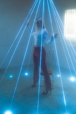 A person with blonde hair stands amidst an array of blue laser beams in a dimly lit room. The clothing consists of a white top and red pants, and the person is wearing high-heeled shoes. A decorative chair is present behind the individual. Light streams in from a window with blinds on the right side.