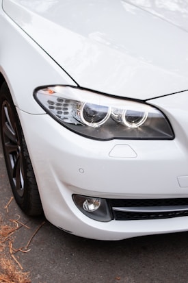 The image captures the front section of a sleek, modern car with a prominent focus on the headlights and grille. The vehicle features a glossy white exterior with a smooth, aerodynamic design. The headlights are LED with circular elements, and the car is parked on a road with some brown leaves scattered on the ground.