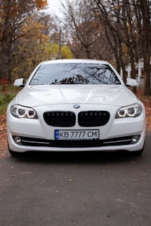 A white BMW car is parked on a paved road surrounded by tall trees with autumn foliage. The front view captures the sleek design of the vehicle, showcasing its headlights and grille. The car has a Ukrainian license plate and the overcast sky is reflected on the windshield.
