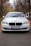 A white BMW car is parked on a paved road surrounded by tall trees with autumn foliage. The front view captures the sleek design of the vehicle, showcasing its headlights and grille. The car has a Ukrainian license plate and the overcast sky is reflected on the windshield.