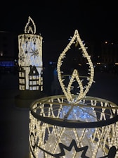 Large decorative candle structures adorned with illuminated lights in a nighttime setting. The candles feature intricate patterns and designs, including snowflakes, and emit a warm glow. The background is dark with faint outlines of buildings and distant lights.