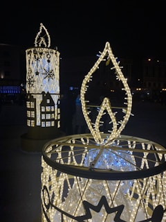 Large decorative candle structures adorned with illuminated lights in a nighttime setting. The candles feature intricate patterns and designs, including snowflakes, and emit a warm glow. The background is dark with faint outlines of buildings and distant lights.