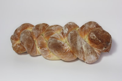 A loaf of braided bread with a golden-brown crust and a dusting of flour on its surface, placed on a plain white background.