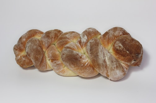 A loaf of braided bread with a golden-brown crust and a dusting of flour on its surface, placed on a plain white background.