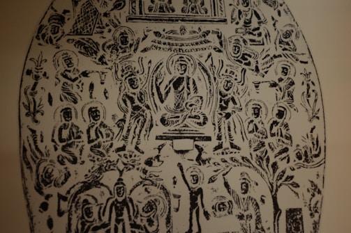 An intricate black and white etching featuring numerous human figures in various seated and standing positions. The central figure appears to be sitting cross-legged, surrounded by an aura, with others arranged symmetrically around them. The figures are stylized and have halos around their heads. Ornate patterns and what appear to be nature elements are interspersed throughout the design.