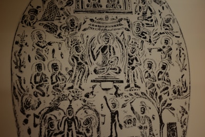 An intricate black and white etching featuring numerous human figures in various seated and standing positions. The central figure appears to be sitting cross-legged, surrounded by an aura, with others arranged symmetrically around them. The figures are stylized and have halos around their heads. Ornate patterns and what appear to be nature elements are interspersed throughout the design.