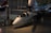 A small, sleek aircraft is positioned inside a spacious hangar. The aircraft features a glossy white exterior with blue and black detailing and is equipped with a large rotor on top, indicating it is likely a gyroplane or rotorcraft. Its reflective surface catches the ambient lighting from the hangar, creating a shiny appearance. In the background, other larger aircraft are partially visible, giving context to the setting as an aviation museum or exhibition space.