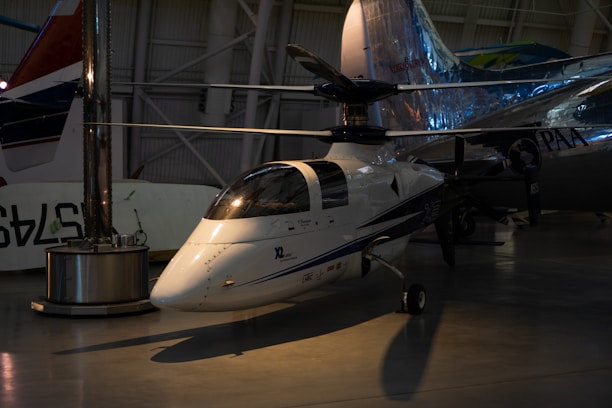 A small, sleek aircraft is positioned inside a spacious hangar. The aircraft features a glossy white exterior with blue and black detailing and is equipped with a large rotor on top, indicating it is likely a gyroplane or rotorcraft. Its reflective surface catches the ambient lighting from the hangar, creating a shiny appearance. In the background, other larger aircraft are partially visible, giving context to the setting as an aviation museum or exhibition space.