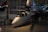 A small, sleek aircraft is positioned inside a spacious hangar. The aircraft features a glossy white exterior with blue and black detailing and is equipped with a large rotor on top, indicating it is likely a gyroplane or rotorcraft. Its reflective surface catches the ambient lighting from the hangar, creating a shiny appearance. In the background, other larger aircraft are partially visible, giving context to the setting as an aviation museum or exhibition space.