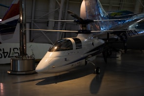 A small, sleek aircraft is positioned inside a spacious hangar. The aircraft features a glossy white exterior with blue and black detailing and is equipped with a large rotor on top, indicating it is likely a gyroplane or rotorcraft. Its reflective surface catches the ambient lighting from the hangar, creating a shiny appearance. In the background, other larger aircraft are partially visible, giving context to the setting as an aviation museum or exhibition space.