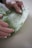 Hands gently holding a piece of green fabric with delicate leaf and flower patterns, featuring bees and geometric shapes. The fabric appears to be soft and possibly made for wrapping or crafting.