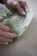 Hands gently holding a piece of green fabric with delicate leaf and flower patterns, featuring bees and geometric shapes. The fabric appears to be soft and possibly made for wrapping or crafting.