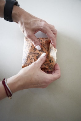 Two hands are wrapping a rectangular object using a piece of fabric with a decorative pattern. The fabric features various floral and leaf designs in earthy tones. The scene is set on a light surface, and the person is wearing a black wristwatch on their left wrist and a red string bracelet on their right wrist.