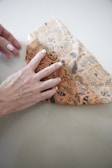 A hand holding and shaping a piece of beige wax paper decorated with black illustrations of bees, honeycombs, and leaves on a light gray surface.