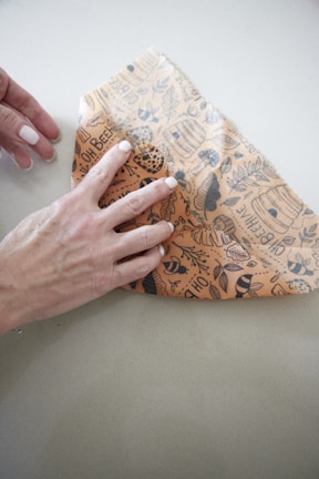 A hand holding and shaping a piece of beige wax paper decorated with black illustrations of bees, honeycombs, and leaves on a light gray surface.