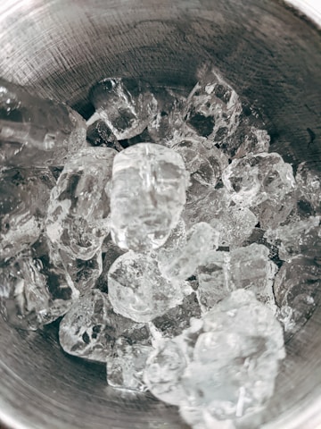 Large chunks of clear ice fill a metal bowl with a textured inner surface. The ice cubes are irregular in shape and densely packed together.