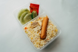 A clear plastic container holds a portion of fried rice, garnished with a sausage and small pieces of chicken. Fresh slices of cucumber and lime are placed beside a packet of sauce in the compartment.