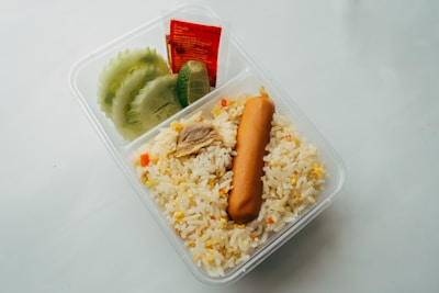 A clear plastic container holds a portion of fried rice, garnished with a sausage and small pieces of chicken. Fresh slices of cucumber and lime are placed beside a packet of sauce in the compartment.
