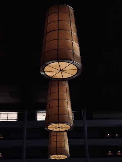 The image displays a dark interior space with large, cylindrical hanging lights. The lights are arranged vertically and appear to be suspended from the ceiling. The background features a multi-story building with silhouetted figures visible through the windows, creating a dramatic contrast against the lit elements.