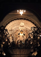 A beautifully crafted statue of a multi-armed deity is showcased in a richly detailed setting. The deity is adorned with intricate designs and is surrounded by smaller figures. The scene is lit by a chandelier above, casting a warm glow over the artwork. Traditional elements and artistic patterns emphasize the cultural significance.
