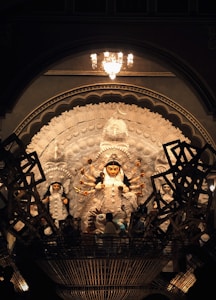A beautifully crafted statue of a multi-armed deity is showcased in a richly detailed setting. The deity is adorned with intricate designs and is surrounded by smaller figures. The scene is lit by a chandelier above, casting a warm glow over the artwork. Traditional elements and artistic patterns emphasize the cultural significance.