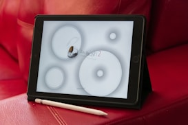 A digital tablet displaying a drawing app interface rests on a red leather couch. An electronic stylus is placed next to the tablet. The app screen shows circular brush tools and the words 'Zen Brush 2' indicating its function as a digital drawing tool.