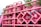 A stack of pink gas cylinders are secured onto a vehicle with a pink metal frame. The cylinders have text on them and appear to be part of a transport or delivery setup. There are some signs of dirt or wear on the cylinders. In the background, there are some green tree leaves visible, adding a natural element to the setting.