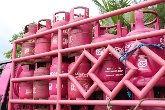 A stack of pink gas cylinders are secured onto a vehicle with a pink metal frame. The cylinders have text on them and appear to be part of a transport or delivery setup. There are some signs of dirt or wear on the cylinders. In the background, there are some green tree leaves visible, adding a natural element to the setting.