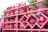 A stack of pink gas cylinders are secured onto a vehicle with a pink metal frame. The cylinders have text on them and appear to be part of a transport or delivery setup. There are some signs of dirt or wear on the cylinders. In the background, there are some green tree leaves visible, adding a natural element to the setting.