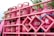 A stack of pink gas cylinders are secured onto a vehicle with a pink metal frame. The cylinders have text on them and appear to be part of a transport or delivery setup. There are some signs of dirt or wear on the cylinders. In the background, there are some green tree leaves visible, adding a natural element to the setting.