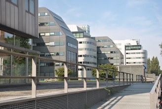 A series of modern office buildings with angular and curved architectural designs, featuring large windows and metallic facades. Surrounding the buildings, there are trees and greenery, and a bridge or walkway with metal railings stretches across the foreground, leading up to the structures. The sky is clear with a light blue hue.