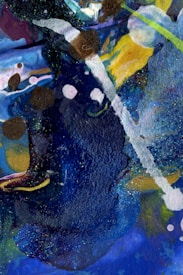 An abstract painting featuring a vibrant mix of colors and textures with bold blue strokes and speckles of glitter. White and brown patches overlap, creating a dynamic contrast against the dark background. Splashes of yellow and hints of green add vibrancy, while fluid shapes create a sense of movement.