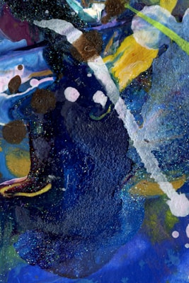 An abstract painting featuring a vibrant mix of colors and textures with bold blue strokes and speckles of glitter. White and brown patches overlap, creating a dynamic contrast against the dark background. Splashes of yellow and hints of green add vibrancy, while fluid shapes create a sense of movement.
