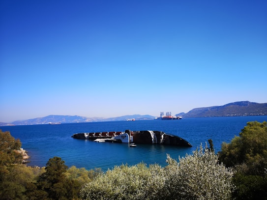 A large fragment of a shipwreck is partially submerged in clear blue water. The backdrop features distant mountains and a bright blue sky without any clouds. In the background, there are additional ships or structures along the horizon. The foreground includes green vegetation and some trees.
