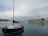 A marina scene with a small sailboat in the foreground, featuring a wooden hull and a white cabin. In the background is a large ferry with the name Levante Ferries and the number 1880 painted on the side. The sky is overcast with scattered clouds, and the water is calm, reflecting the boats.