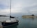 A marina scene with a small sailboat in the foreground, featuring a wooden hull and a white cabin. In the background is a large ferry with the name Levante Ferries and the number 1880 painted on the side. The sky is overcast with scattered clouds, and the water is calm, reflecting the boats.