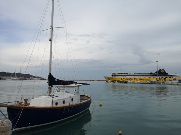 A marina scene with a small sailboat in the foreground, featuring a wooden hull and a white cabin. In the background is a large ferry with the name Levante Ferries and the number 1880 painted on the side. The sky is overcast with scattered clouds, and the water is calm, reflecting the boats.