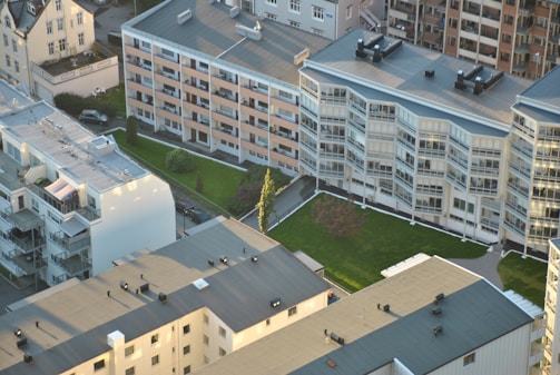 Aerial view of a residential area featuring several multi-story apartment buildings with flat roofs, surrounded by green lawns and pathways. The buildings have large windows, balconies, and varied architectural designs. Vehicles and roads are visible, emphasizing urban living.