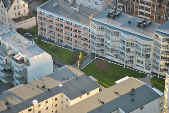Aerial view of a residential area featuring several multi-story apartment buildings with flat roofs, surrounded by green lawns and pathways. The buildings have large windows, balconies, and varied architectural designs. Vehicles and roads are visible, emphasizing urban living.