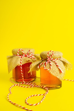 Two glass jars filled with a light amber liquid are topped with rustic burlap covers, tied with red and white striped twine. These jars are set against a bright yellow background, and one jar has a loose length of twine trailing across the surface.