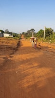 A rural dirt road stretches into the distance under a clear blue sky. Several people, including women in traditional clothing carrying items on their heads, walk along the road. The area is lush with green trees and vegetation lining the edges of the road, while utility poles follow the path. A white building is visible on the left, contrasting with the natural surroundings.