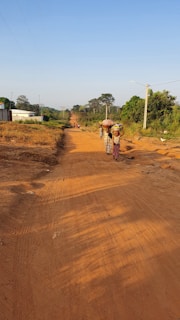 A rural dirt road stretches into the distance under a clear blue sky. Several people, including women in traditional clothing carrying items on their heads, walk along the road. The area is lush with green trees and vegetation lining the edges of the road, while utility poles follow the path. A white building is visible on the left, contrasting with the natural surroundings.